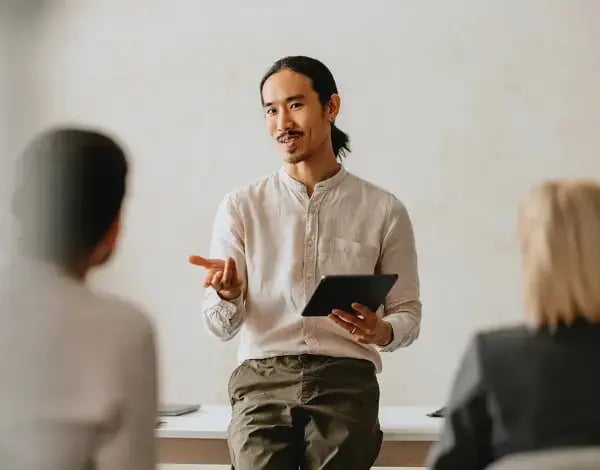 Photo of an Asian man holding a tablet and speaking to two people sat in front of him