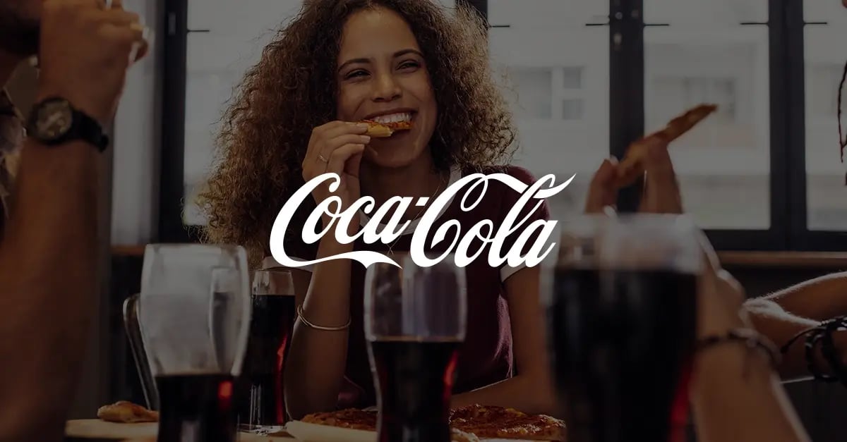 Photo of a group of people sat around a table eating pizza with a white version of the Coca Cola logo superimposed in the middle foreground