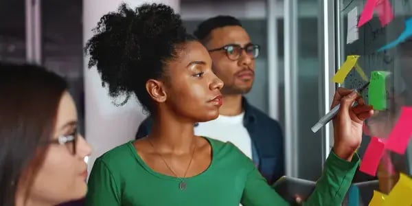 Two women and a man stood in front of a glass wall with post-it notes stuck to it and one woman writing something on one of them